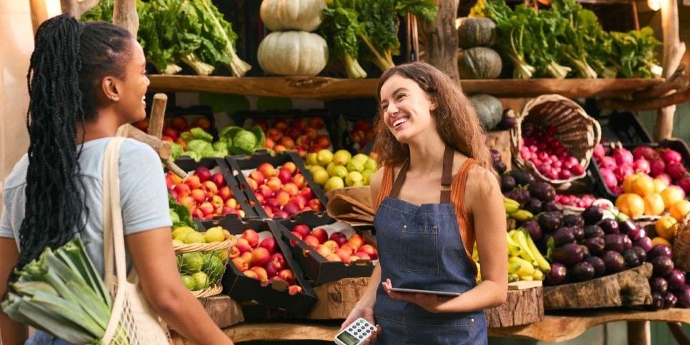 A smiling worker in a denim apron assisting a customer shopping for fresh vegetables and fruit at an open-air community market stall.